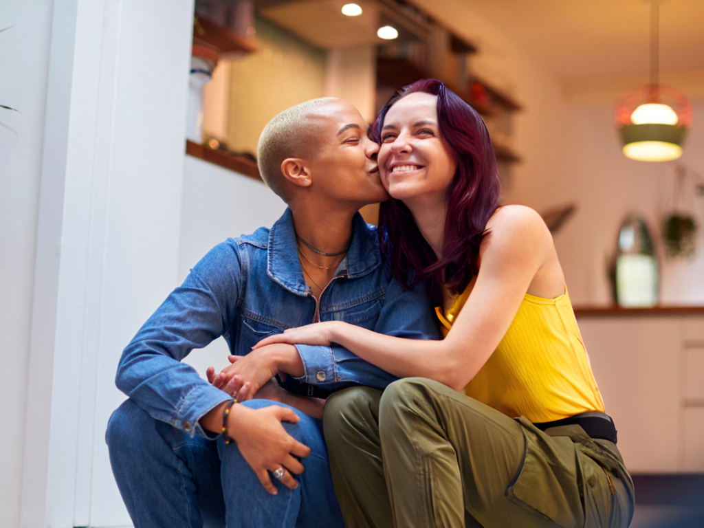 a young couple, a woman short blond hair and medium brown skin and woman with red hair and light skin, sits in the kitchen of their home protected with a homeowners insurance policy