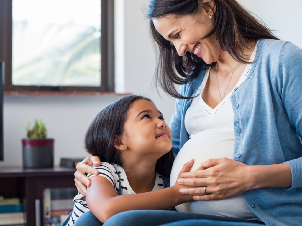 a pregnant mother with medium brown skin, dark hair, and blue sweater holds the hand of her daughter on her belly, a girl with brown skin, dark hair, and striped shirt as she remembers why annual insurance reviews are important during life changes