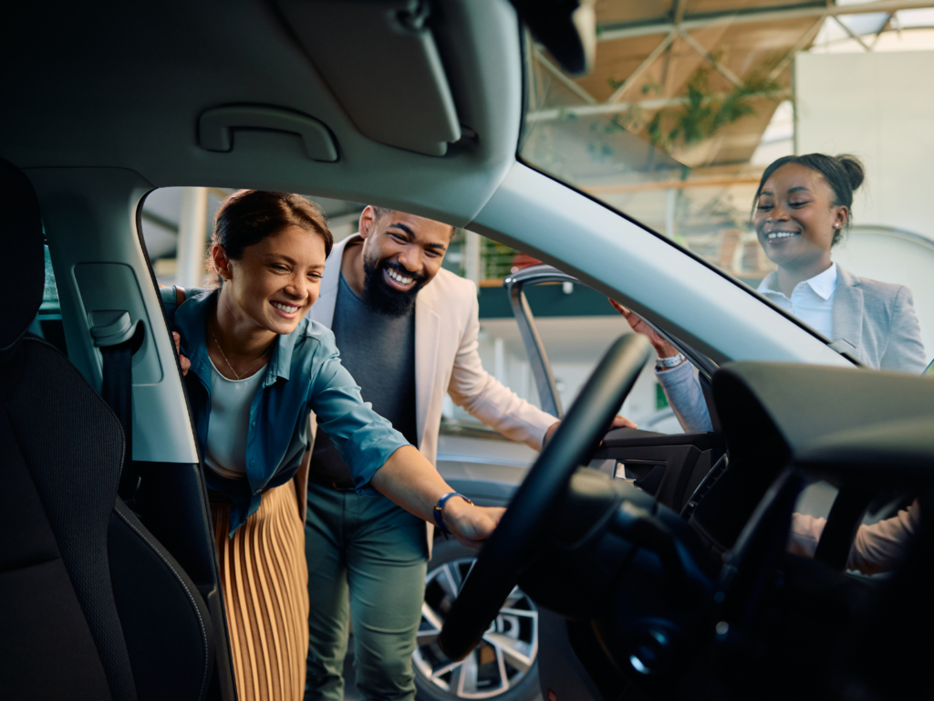 a woman with pale skin and yellow skirt with a man with dark brown skin and dark hair in a blazer look inside a car they are considering purchasing with the salesperson